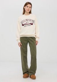 Felpa color crema con grafica "YELLOWSTONE National Park" e design di montagne, abbinata a pantaloni larghi verde oliva e pantofole marroni.