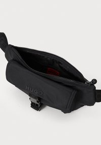 Sac banane noir en nylon avec fermeture éclair, doté d'un logo à l'avant et d'une sangle à boucle. L'intérieur a une doublure lisse.