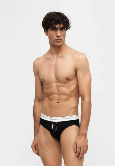 Homme aux cheveux bouclés et foncés portant un slip noir avec une ceinture blanche, debout devant un fond clair uni.