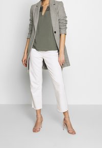 Blazer lungo grigio su una blusa verde con scollatura in pizzo, abbinato a jeans bianchi a vita alta e tacchi nudi con cinturini.