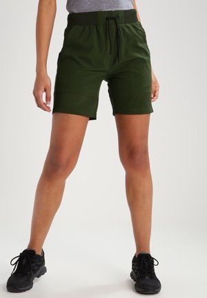 Persoon draagt olijfgroene sportshorts met geperforeerde onderste panelen en zwarte hardloopschoenen, staat met gespreide benen tegen een witte achtergrond.