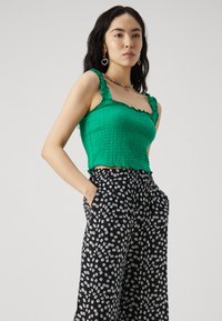 Zelený crop top so smockovaním a volánovými ramienkami, skombinovaný s čiernymi širokými nohavicami s bielym potlačou harmančeka. Hladká textúra, priliehavý strih.