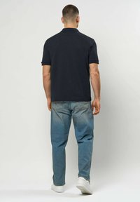 Mann som går bortover iført svart kortermet poloskorte, falmede blå jeans og hvite joggesko mot en ensfarget bakgrunn.