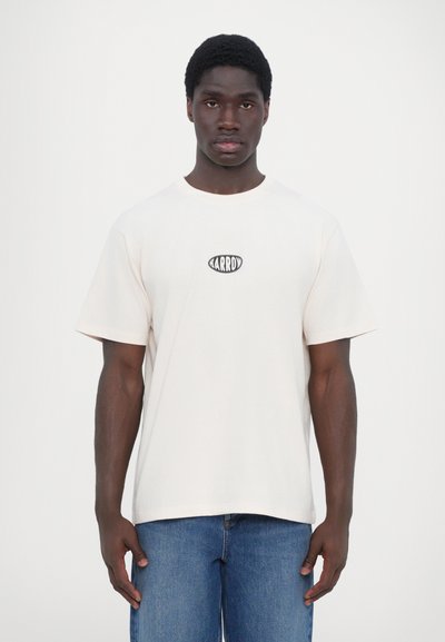 T-shirt en coton couleur crème à manches courtes, col rond et avec un petit logo ovale noir sur la poitrine. Associé à un jean bleu.
