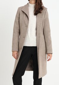Beige wool coat with a notched collar, front button closure, and structured fit. Worn over a light gray sweater and black pants.