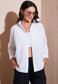Camicia bianca con bottoni realizzata in tessuto leggero, con una vestibilità rilassata e maniche arrotolate, abbinata a un top bianco corto e pantaloni scuri.