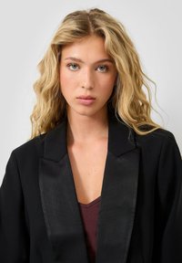 Jauna sieviete ar gariem viļņainiem blondiem matiem, tērpusies melnā blazerī un tumšā augšpusē, skatās tieši kamerā ar neitrālu izteiksmi.