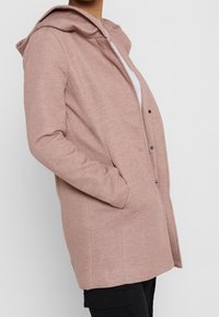 Mujer vestida con un abrigo de lana rosa claro con capucha y bolsillos frontales, parcialmente abotonado sobre una blusa blanca y pantalones negros, de pie de lado.