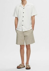 Man wearing a short-sleeve white button-up shirt, beige knee-length shorts, and tan open-toe sandals with straps.