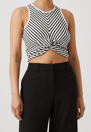 Osoba ubrana w czarno-biały bezrękawnik w paski z przodu skręcony i czarne spodnie z wysokim stanem, odsłaniająca brzuch i tatuaż na ramieniu.