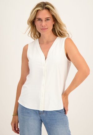 Blondine vrouw draagt een mouwloze witte blouse met knopen en blauwe spijkerbroek, staat met één hand op de heup tegen een witte achtergrond.
