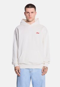 Biała bluza z kapturem i przednią kieszenią oraz czerwonym logo "Frost". Miękka tkanina, luźny krój i regulowany kaptur ze sznurkiem. Noszona z jasnoniebieskimi dżinsami.