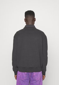 Dark gray sweatshirt with a collar, drop shoulders, and ribbed cuffs. The fabric appears soft and relaxed, paired with bright purple jeans.