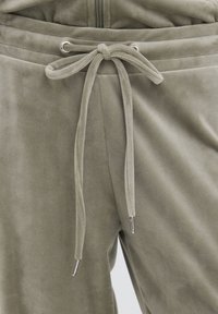 Pantaloni in velluto morbido verde grigio con coulisse regolabile annodata a fiocco sulla fascia elastica in vita e una cerniera parziale visibile sopra.
