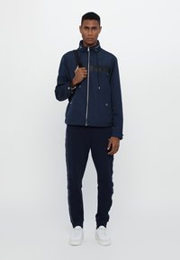 Marine vanntett jakke med glidelås foran, snøring på hetten og svart brystdetalj. Kombinert med matchende joggebukser og hvite sneakers.