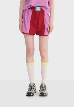 Osoba ubrana w czerwone i różowe szorty z niebieską metką, białe skarpety do kolan z żółtymi paskami oraz szare buty do biegania z czarnymi sznurówkami.