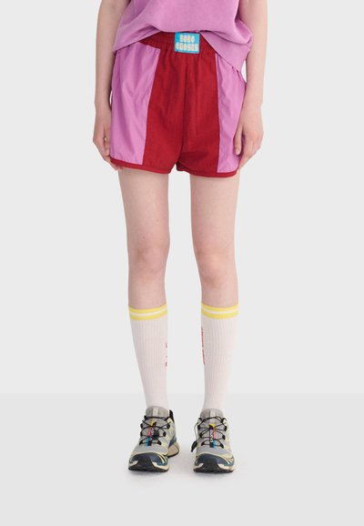 Personne portant un short rouge et rose avec une étiquette bleue, des chaussettes blanches montantes à rayures jaunes, et des chaussures de course grises avec des lacets noirs.