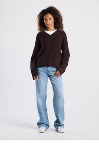 Ruskea neulottu V-kaula-aukollinen ja pitkähihainen paita, yhdistetty vaaleansinisiin, leveälahkeisiin farkkuihin ja valkoisiin tennareihin. Tasainen, teksturoitu pinta.