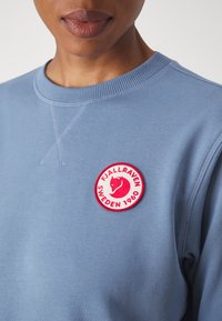 Helles blaues Sweatshirt mit einem runden roten und weißen Patch, das ein Fuchs-Logo und den Text "FJÄLLRÄVEN SCHWEDEN 1960" auf der Brust gestickt hat.