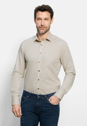 Man met kort donker haar en baard, die een beige overhemd met knopen en een blauwe spijkerbroek draagt, staat met één hand in de zak.