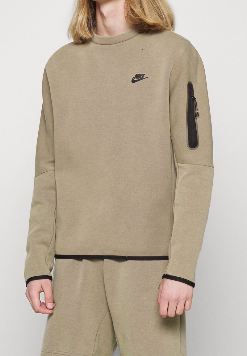 Osoba v béžové mikině Nike a odpovídajících kalhotách, s černým logem Nike na hrudi a zipovou kapsou na levém rukávu.