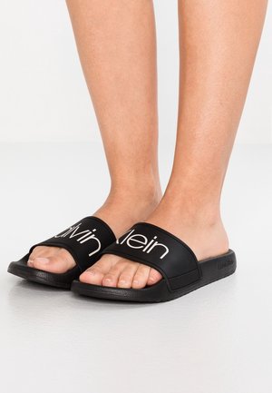 Stopy w czarnych klapkach Calvin Klein z białym logo na paskach, stojące na jasnoszarej podłodze.