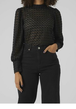 Kvinde iført en gennemsigtig sort langærmet mønstret bluse stoppet ned i sorte højtaljede bukser, hånd i lommen på en grå baggrund.