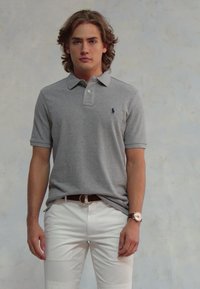 Jaunas vyras su vidutinio ilgio plaukais, vilkintis pilką polo marškinėlį ir baltas kelnes, stovi prieš paprastą šviesią sieną.