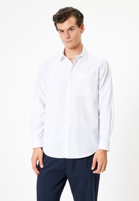 Chemise blanche à boutons avec une poche, manches longues, col classique et texture lisse, associée à un pantalon bleu marine foncé.