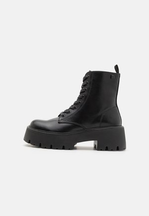 Botas de cuero negro hasta el tobillo con una suela gruesa, punta redonda y cordones, que cuentan con una tira en la parte trasera y suela texturizada.