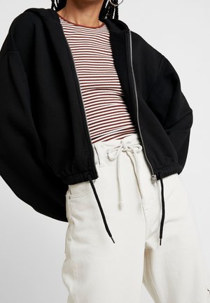 Zwarte rits-hoodie met elastische zoom, gedragen over een rood-wit gestreept shirt en witte hoge taille broek met koordsluiting.