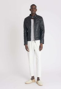 Veste en cuir noir de moto avec poches zippées à l'avant, portée sur une chemise grise, associée à un pantalon blanc cintré et des chaussures slip-on beige clair.