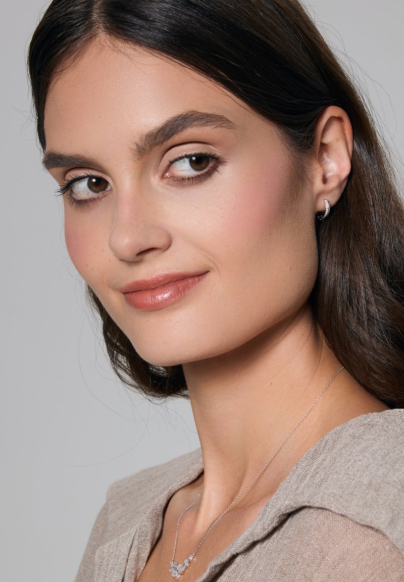 Modelka o brązowej skórze, nosząca srebrny naszyjnik z okrągłymi akcentami i dopasowane kolczyki wkrętki, ubrana w beżową teksturowaną bluzkę.
