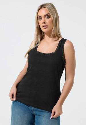 Lyshåret kvinde iført sort ærmeløs top med blonder og blå jeans, poserer foran en ensfarvet lys baggrund.