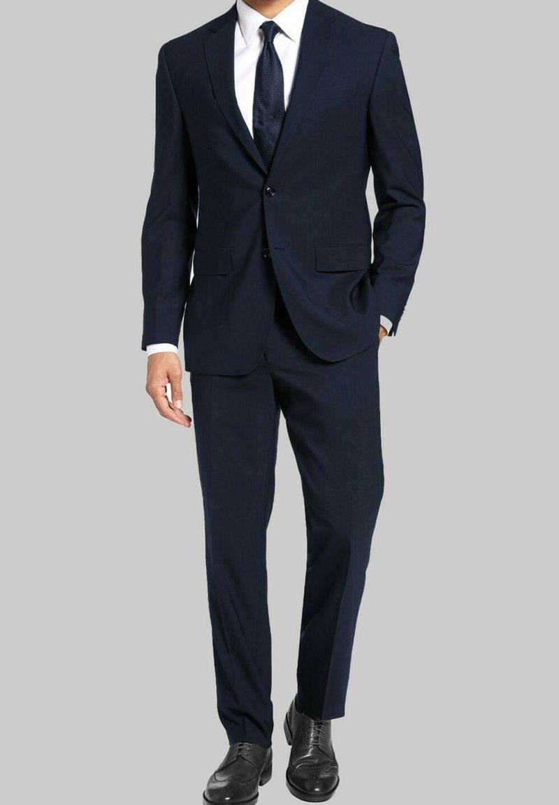 Costume bleu marine avec une fermeture à un bouton, des revers crantés et une coupe ajustée. Chemise blanche et cravate texturée, complétées par des chaussures noires.