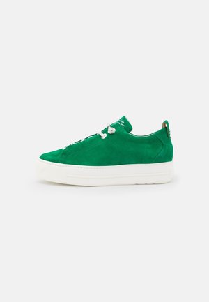 Zielony zamszowy niski sneaker z białą grubą podeszwą i białymi sznurowadłami, widok z boku na jednolite jasnoszare tło.