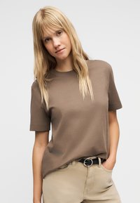 Νεαρή γυναίκα με μακριά ξανθά μαλλιά στέκεται με το ένα χέρι στην τσέπη, φορώντας καφέ πουκάμισο με κοντά μανίκια και μπεζ παντελόνι με μαύρη ζώνη.