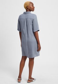 Graues Leinenkurzes Kleid mit Kragen, hochgekrempelten Ärmeln und Rückenschlitzdetails, entworfen für eine lässige Passform. Silberne Sandalen getragen.
