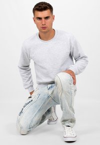 Felpa grigia con maniche lunghe, abbinata a jeans chiari strappati con cuciture decorative e logo ricamato. Sneakers bianche.