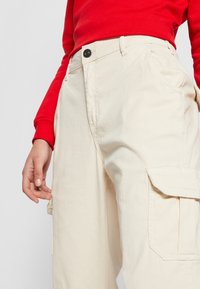Beige cargo pants with a textured fabric, featuring a side pocket and a black button at the waist. The top is a red long-sleeve shirt.