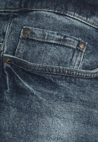 Los jeans de mezclilla azul oscuro presentan un bolsillo trasero con botones en tono plateado y costuras en contraste, destacando una sutil textura desgastada.