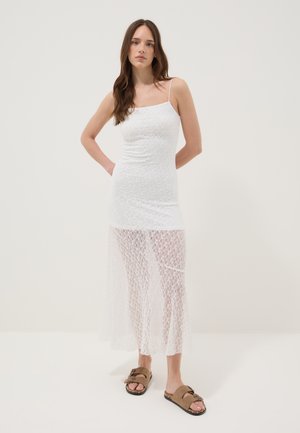 Mujer con cabello largo que lleva un vestido sin mangas de encaje blanco con panel de falda transparente y sandalias abiertas de punta con hebilla color beige, de pie con las manos detrás de la espalda.