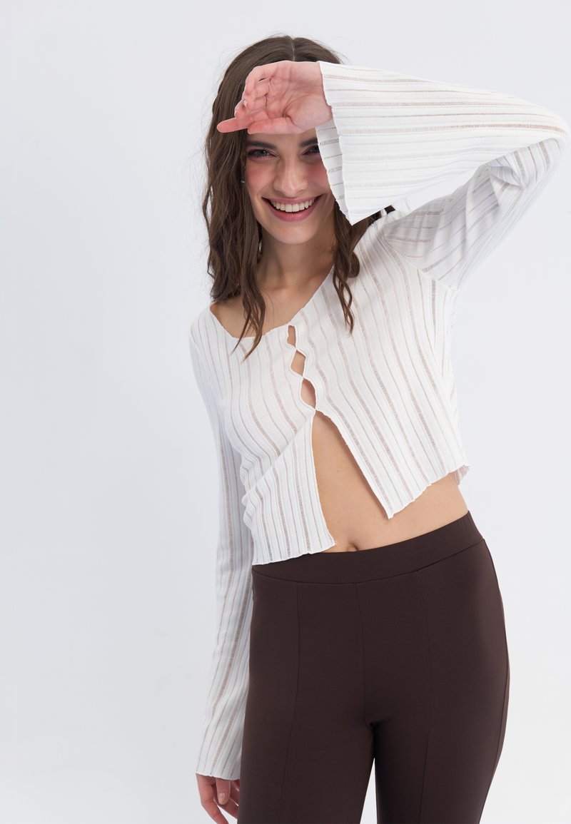Fehér, bordázott crop top, vállnélküli kialakítással, hosszú, bővülő ujjakkal és elöl hasított résszel, magas derekú barna leggingshez párosítva.