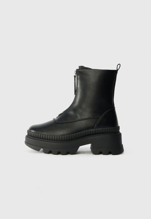 Bota de tornozelo em couro preto com fecho de zip, sola de borracha grossa, detalhes ribulados à volta da sola e uma laço na parte de trás.