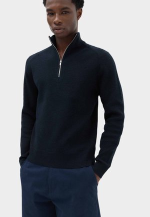 Ung mand iført en sort halv-lynlås sweater og mørkeblå bukser med hænderne i lommerne mod en ensfarvet lys baggrund.