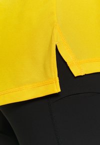 Żółty sportowy top z teksturowanej tkaniny, z bocznymi rozcięciami i kontrastującym czarnym materiałem poniżej. Widoczne przeszycia wzdłuż krawędzi.