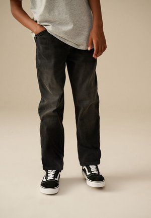 Personne portant un jean noir délavé, des baskets noires et blanches, et un t-shirt gris, debout avec une main dans la poche devant un fond beige.