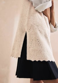 Jasno beżowy sweter z dzianiny o fakturowanym wzorze w zygzak i falbaniastym wykończeniu, w zestawieniu z czarną spódnicą z bocznym rozcięciem.