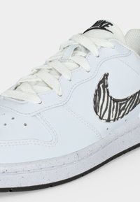 Λευκό αθλητικό παπούτσι Nike με λευκά κορδόνια, ασπρόμαυρο μοτίβο ζέβρας στο Nike swoosh και μαύρο λογότυπο Nike στη γλώσσα.