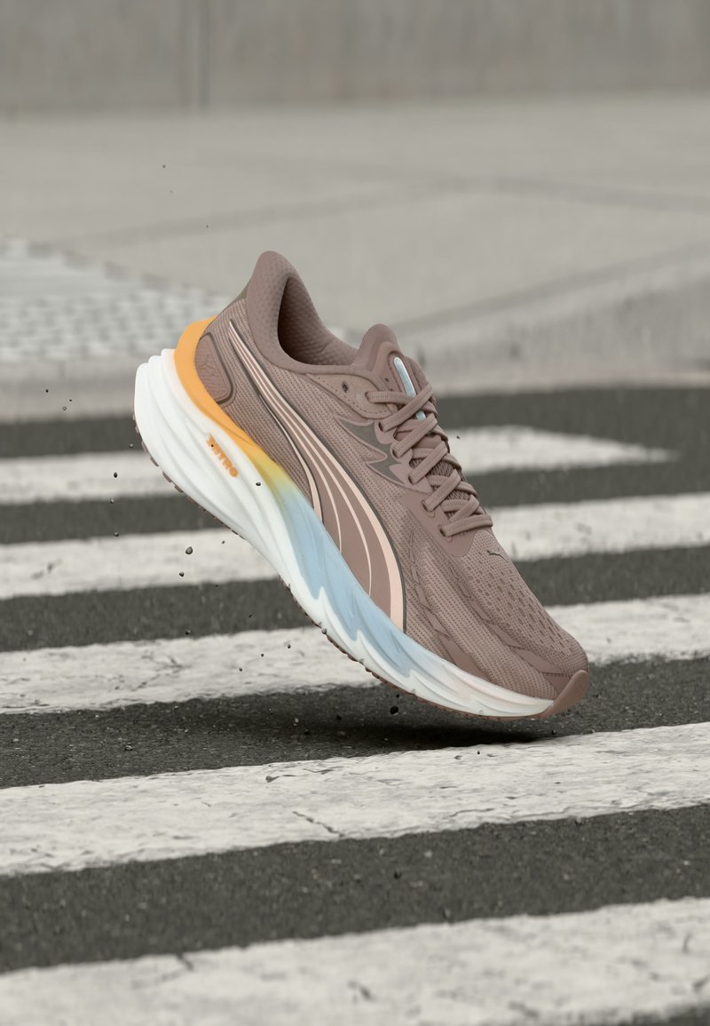 Καφέ αθλητικό παπούτσι με λευκή, μπλε και κίτρινη σόλα στον αέρα πάνω από μια ασπρόμαυρη ραβδωτή διάβαση πεζών σε εξωτερικό χώρο.